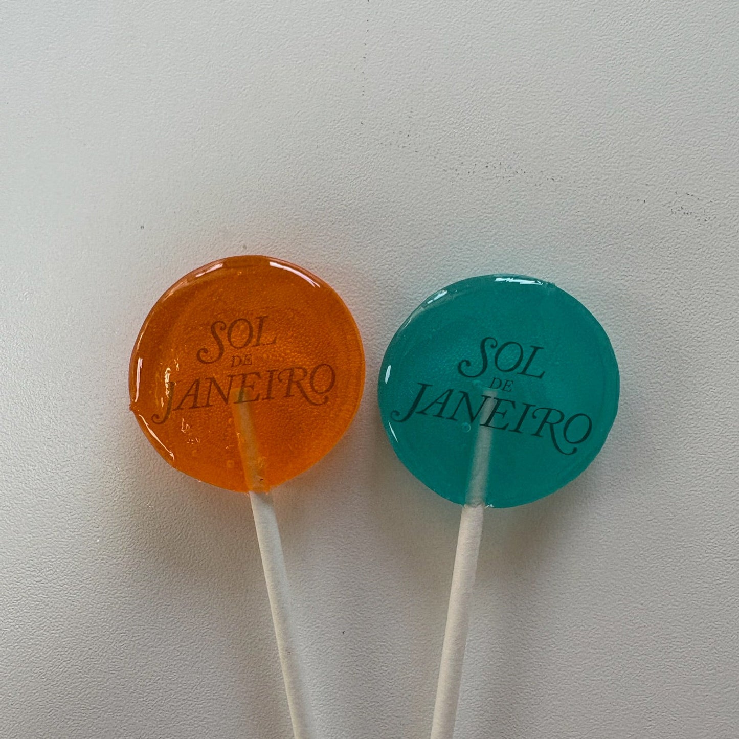 Two lollipops, one orange and one blue, on a white background with 'Sol Janeiro' branding.