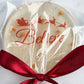 A round holiday lollipop in clear cellophane wrapping with red 'Believe' script and Santa sleigh graphics, tied with a dark red ribbon on a white stick, set on a light marble surface.