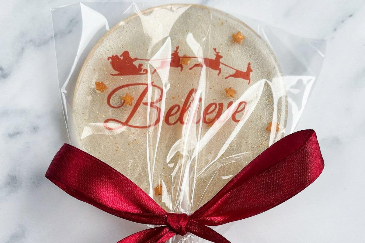 A round holiday lollipop in clear cellophane wrapping with red 'Believe' script and Santa sleigh graphics, tied with a dark red ribbon on a white stick, set on a light marble surface.
