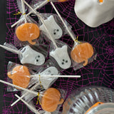 Halloween-themed lollipop treats shaped like orange pumpkins and white ghosts, wrapped individually in clear cellophane on wooden sticks, arranged on a black surface with a purple spiderweb pattern and a white ghost decoration.