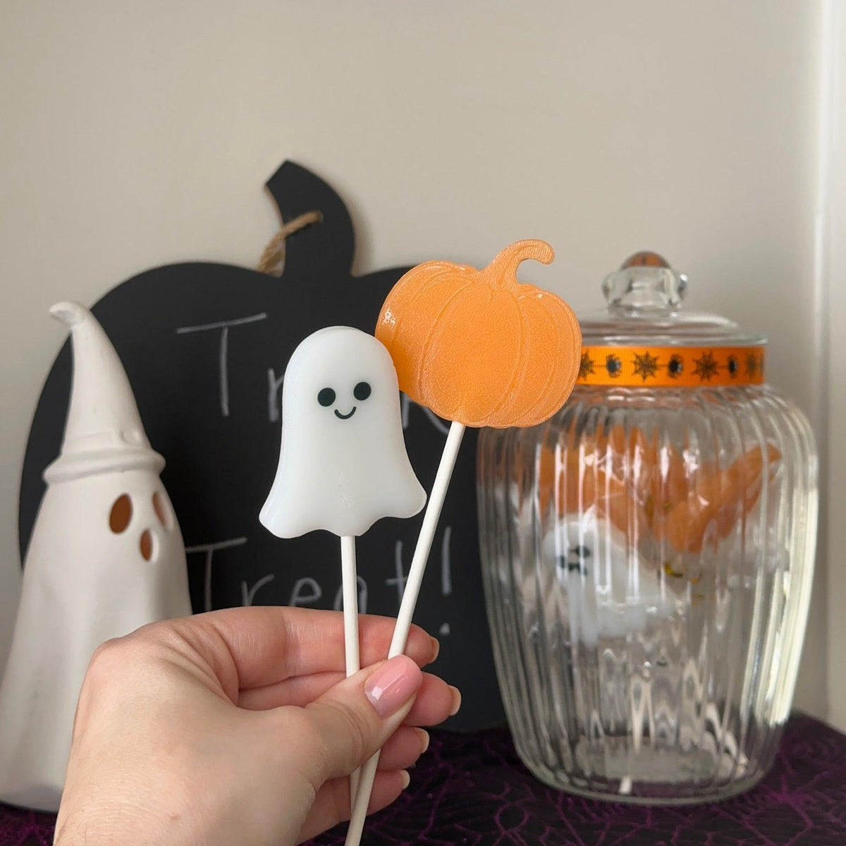 A hand holds two Halloween-themed lollipops: a white ghost-shaped candy on a stick and an orange pumpkin-shaped candy on a stick, with a glass jar and decorative Halloween props in the background.