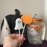 A hand holds two Halloween-themed lollipops: a white ghost-shaped candy on a stick and an orange pumpkin-shaped candy on a stick, with a glass jar and decorative Halloween props in the background.